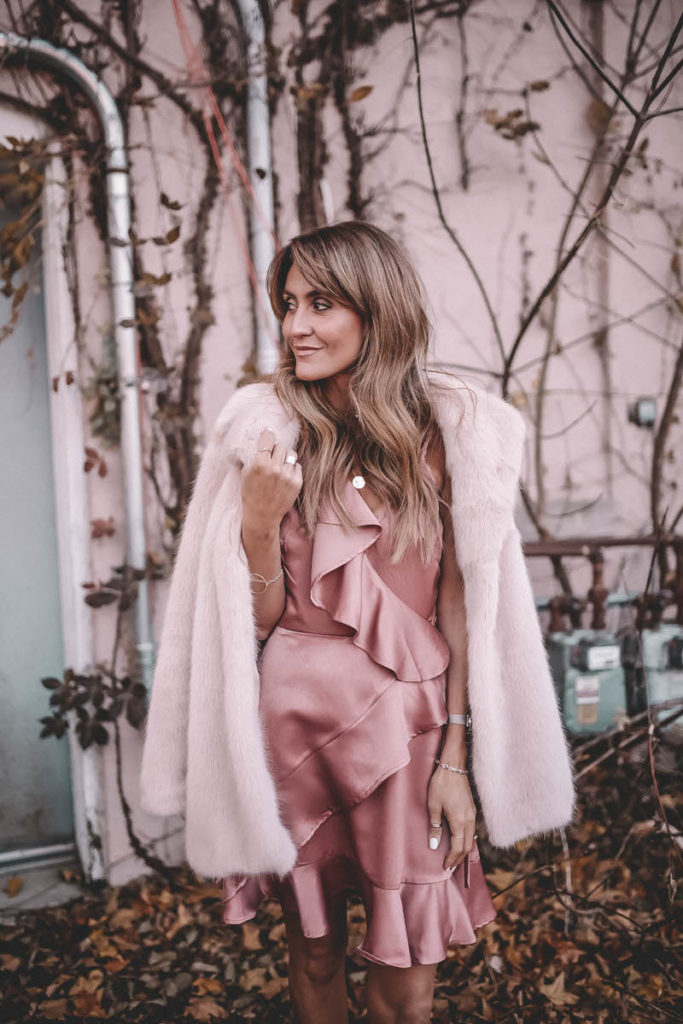 Hottest Pink Holiday Dress featured by top Indianapolis fashion blog, Karina Style Diaries: woman walking in the street wearing a BB Dakota pink ruffle dress, ASOS rose gold sandals, Daniel Wellington watch, grey suede clutch, gold disc necklace and a pink H&M Faux Fur coat